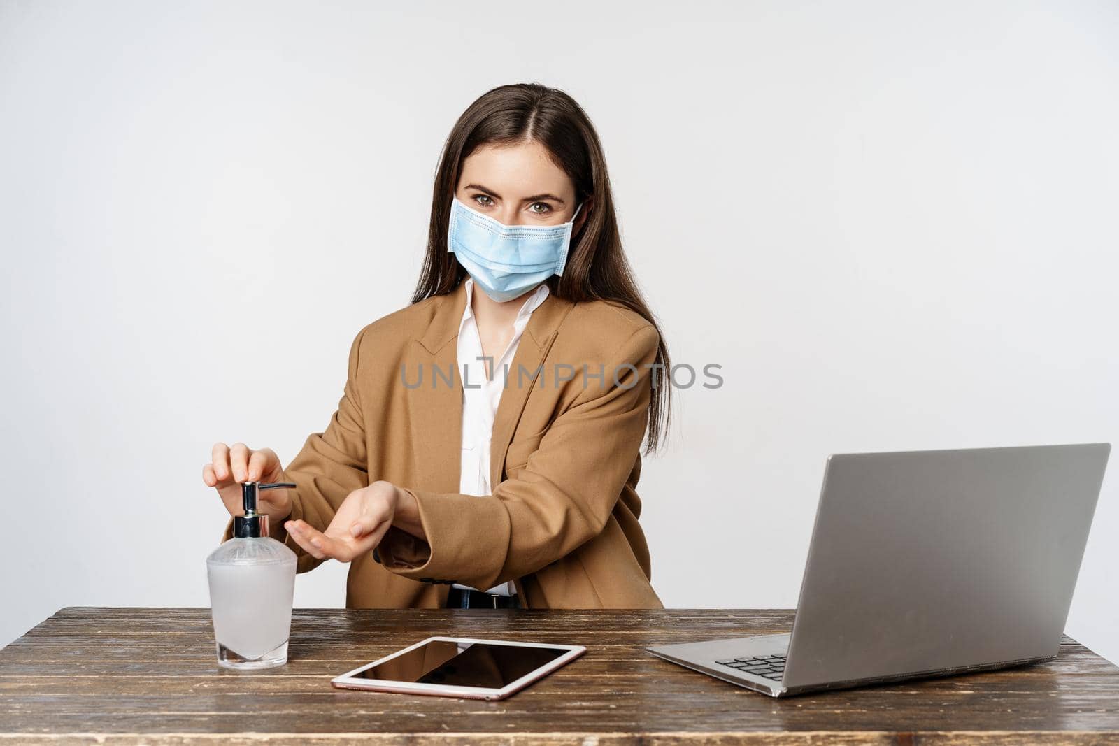 Businesswoman at workplace wearing face mask from covid-19, work in office during pandemic, using hand sanitizer to clean hands, posing over white background.