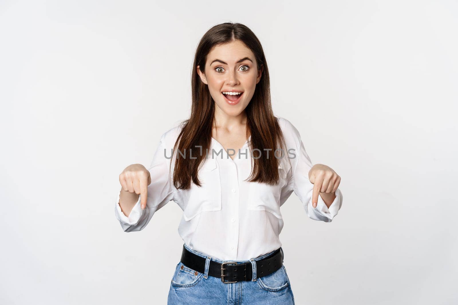 Amazed young woman pointing fingers down, showing announcement or logo banner, looking surprised and intrigued, standing over white background.