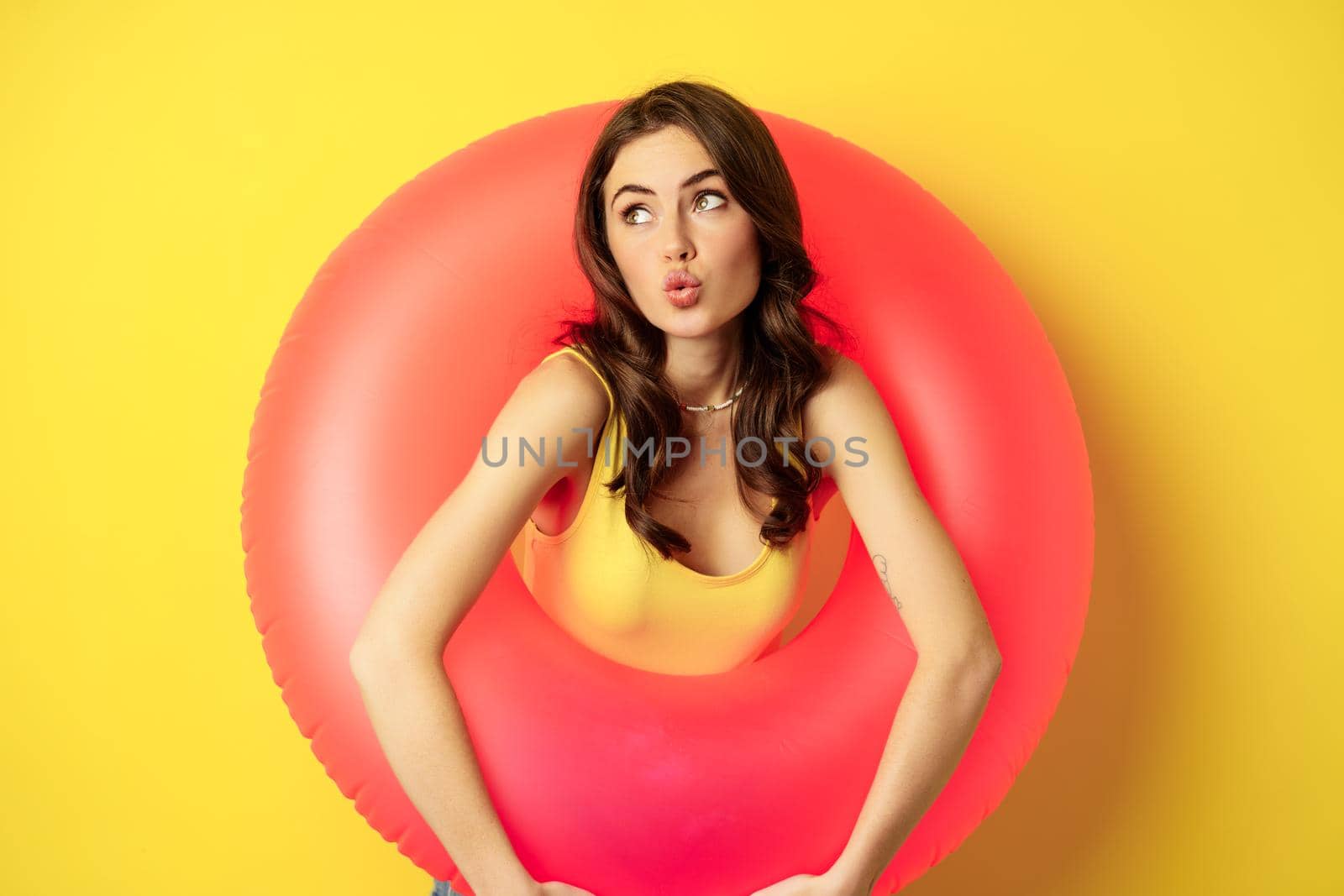 Close up portrait of coquettish young sexy woman inside pink swimming ring, concept of beach holiday, vacation and summer, yellow background.