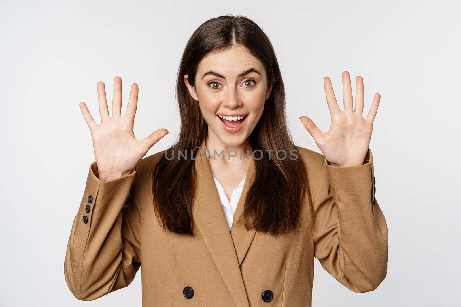 Enthusiastic businesswoman in suit, raising hands palms up, number ten dozen gesture and smiling, standing over white background.