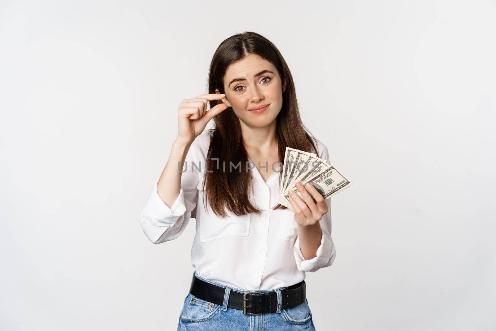 Sad girl pinching fingers and holding money, disappointed in amount of cash, lacking income, standing over white background.
