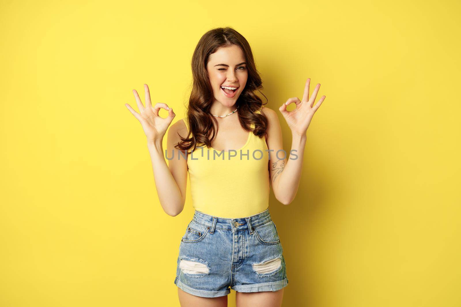 Smiling attractive woman showing okay signs and winking, assure you, praise and compliment smth good, recommending, standing over yellow background.