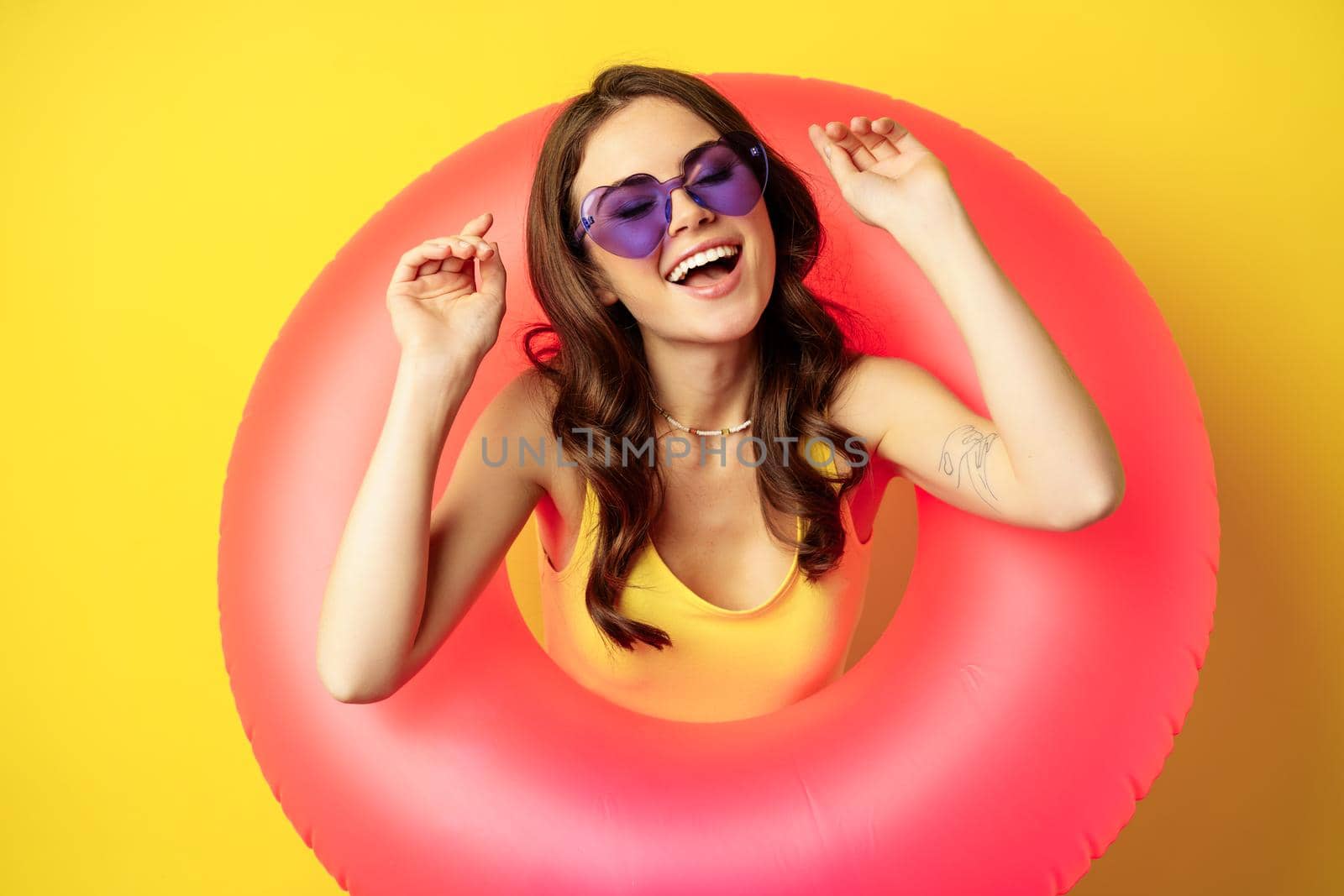 Beautiful happy woman enjoying summer vacation, wearing pink swimming ring on beach, laughing and smiling, standing over yellow background.