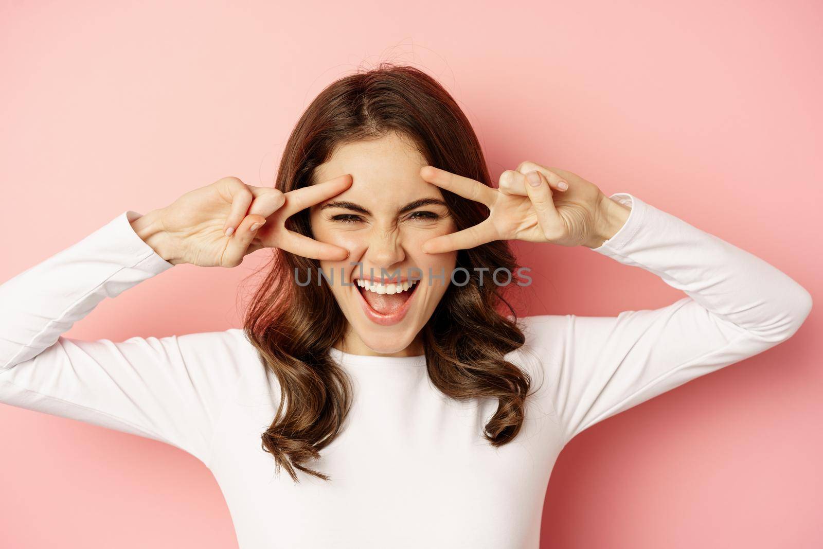 Close up face of young stylish girl, sassy woman showing happy, excited face expression, peace v-sign over eyes, standing against pink background.