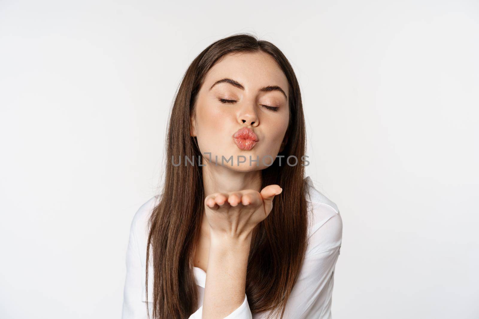 Feminine woman sending air kiss at camera, coquettish flirty pose, kissing, standing over white background. Copy space