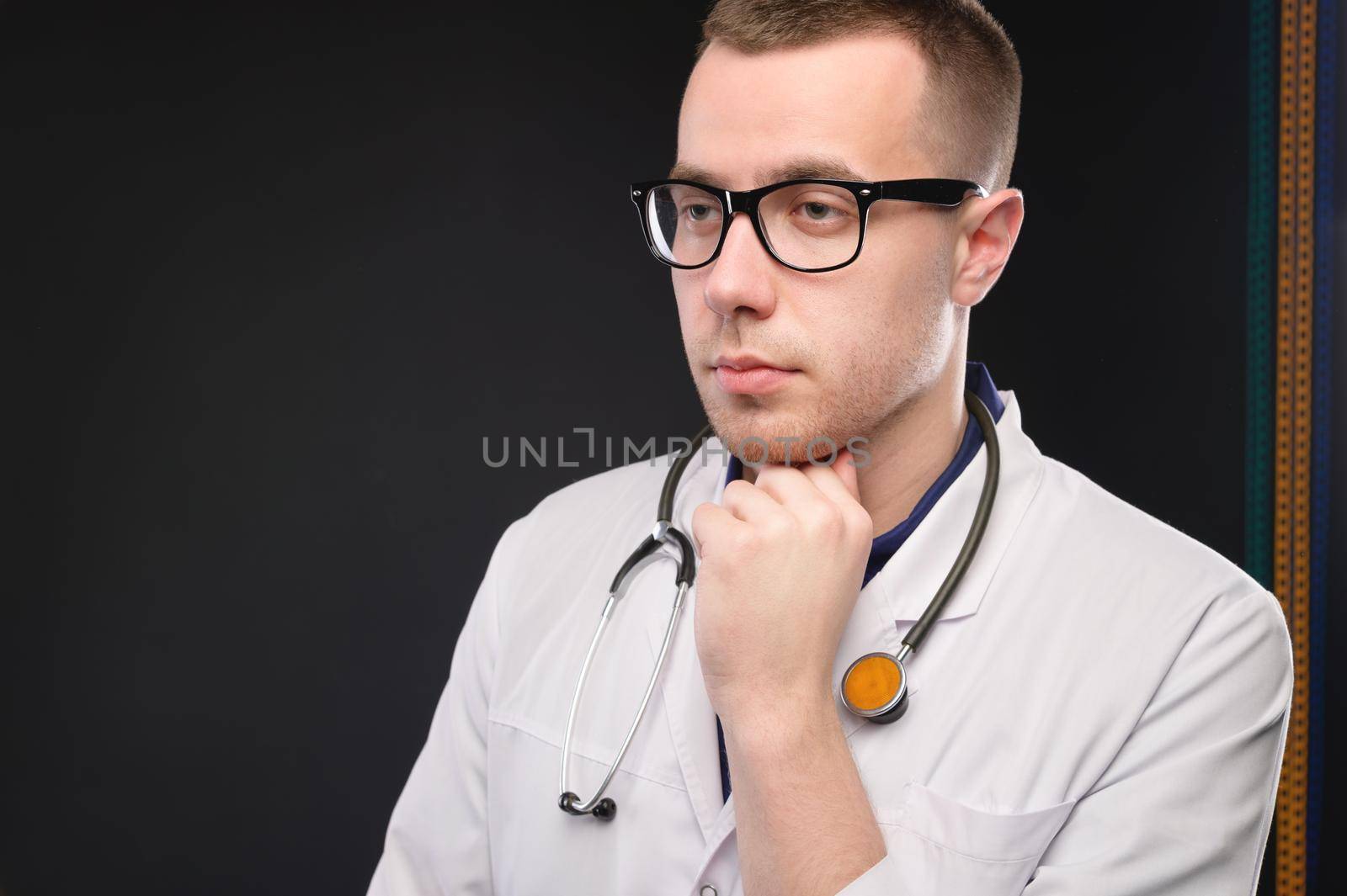 A young attractive Caucasian doctor in glasses and a white coat stands pensive and looks to the side. Studio portrait of a paramedic.