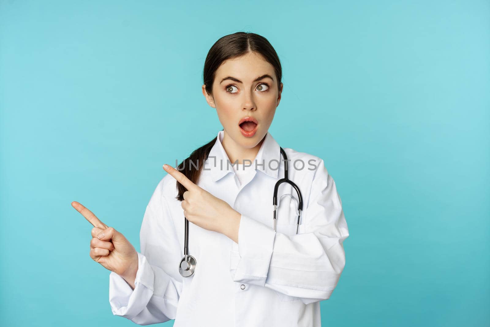 Surprised female doctor, physician with stethoscope, pointing and looking left with amazed, wow face, standing over torquoise background.