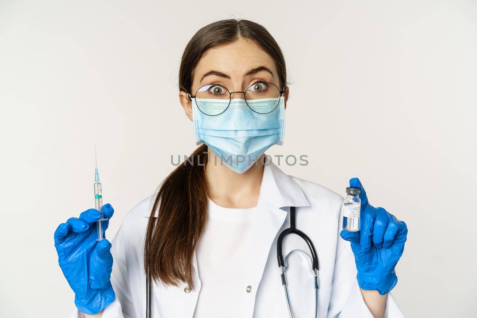 Covid-19 vaccination and healthcare concept. Young doctor in medical mask showing syringe and vaccine from coronavirus omicron variant, standing over white background.