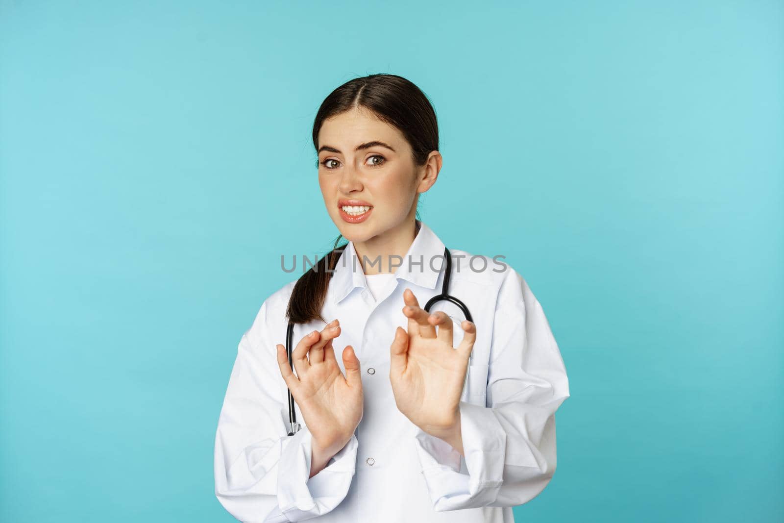 Image of woman doctor cringe, looking with dislike or aversion, rejecting, saying no, stay away, step back from something ugly, standing over torquoise background.