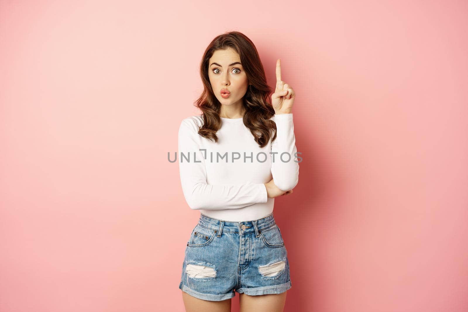 Young brunette glam girl raising finger, pointing up, showing promo offer, eureka gesture, realise something, standing over pink background.