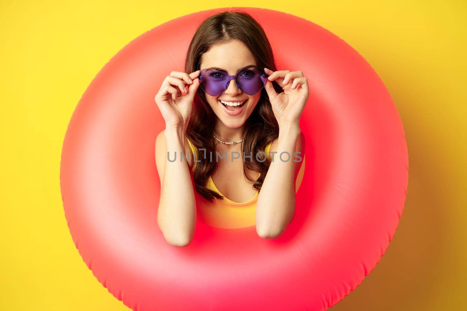Close up portrait of stylish young woman, girl with swimming ring and summer sunglasses, ready for beach holiday, vacation, standing over yellow background.