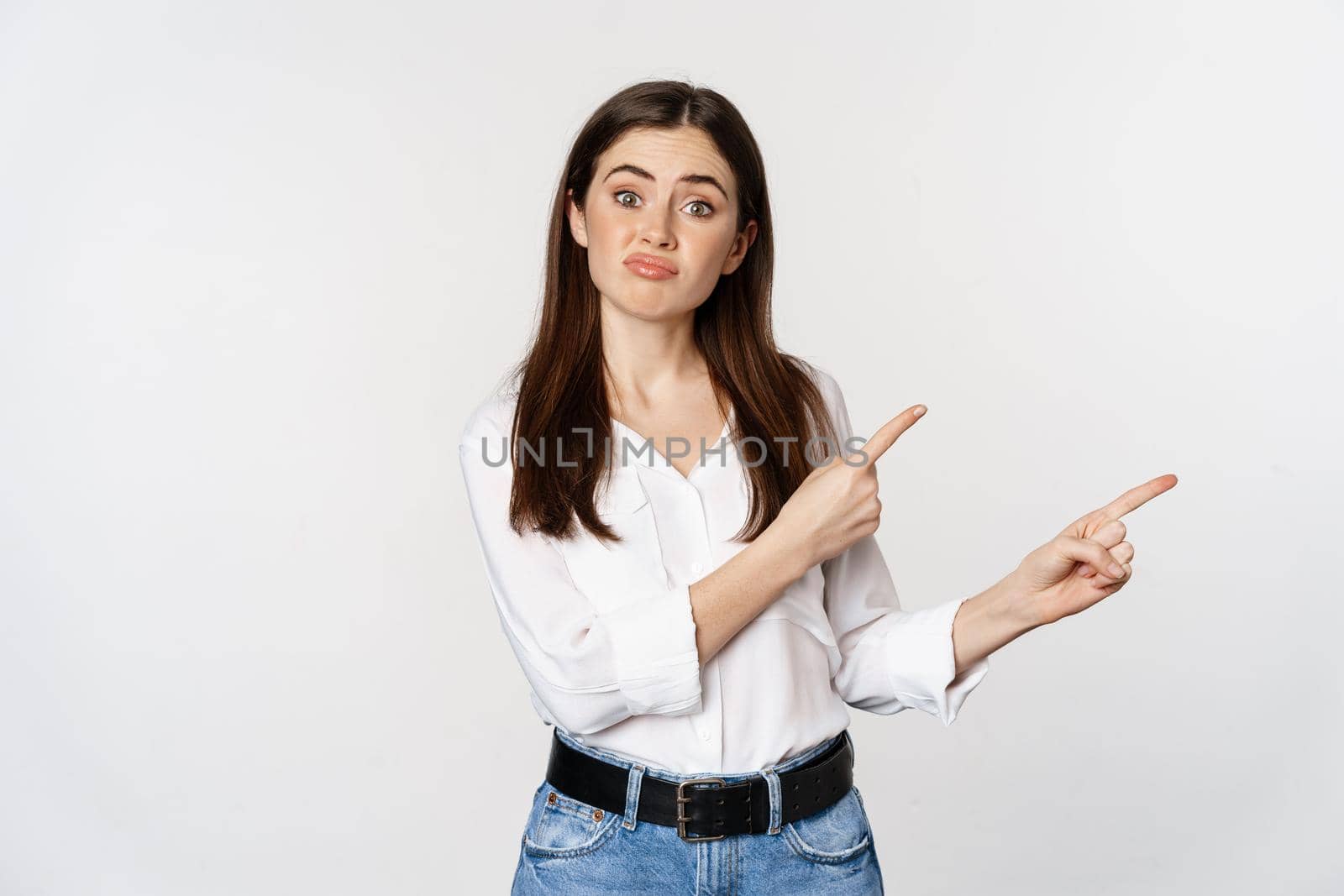 Sad and disappointed cute woman, gloomy girl pointing fingers at logo, complaining, sulking upset, standing in blouse over white background.