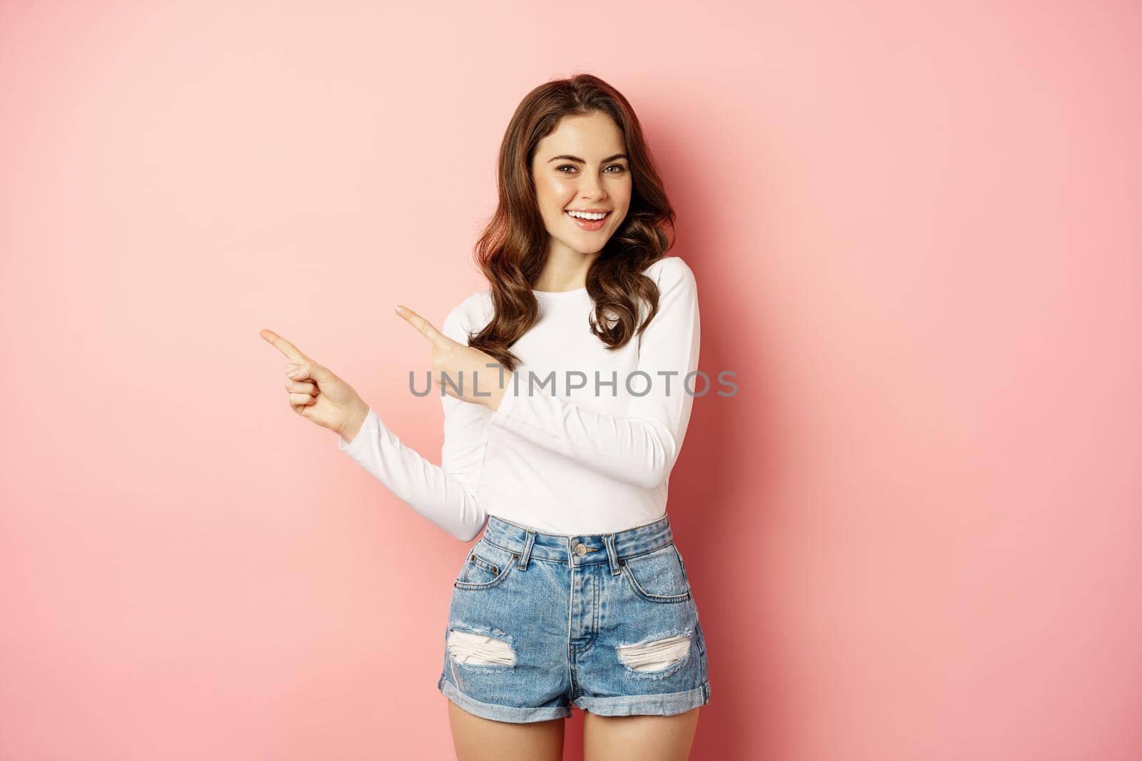 Cute smiling brunette girl, showing advertisement, pointing fingers left at banner, logo on empty pink space, studio background.