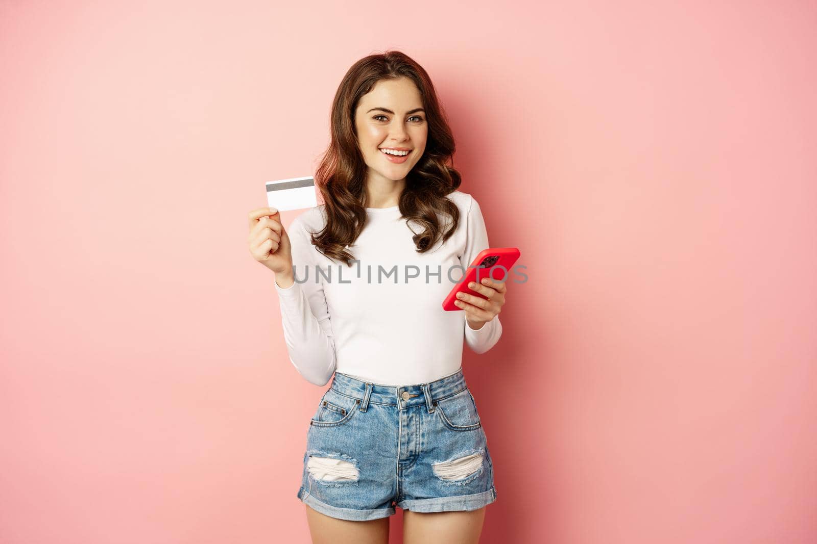 Enthusiastic brunette girl showing credit card and using mobile phone to order or pay, online shopping app, standing over pink background.