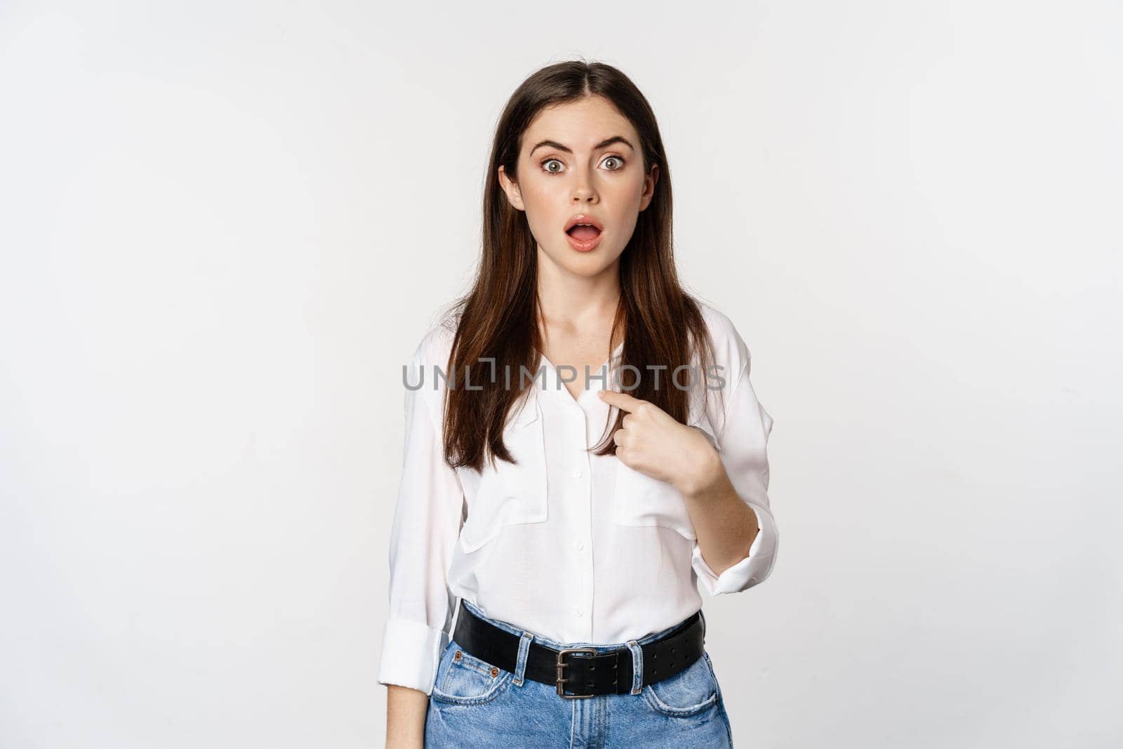 Portrait of young woman pointing at herself with disbelief, surprised face expression, being named or chosen, standing over white background.