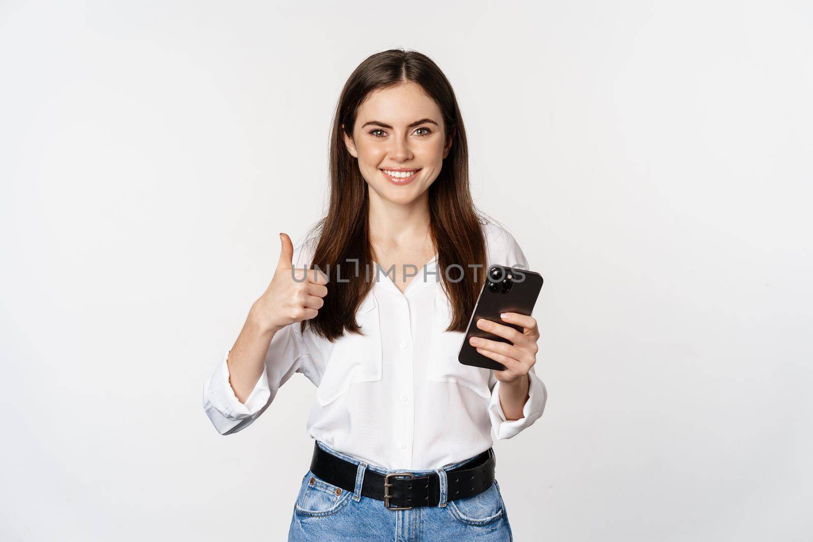 Happy smiling corporate woman, female model showing thumb up, holding smartphone, using cellphone, standing over white background.