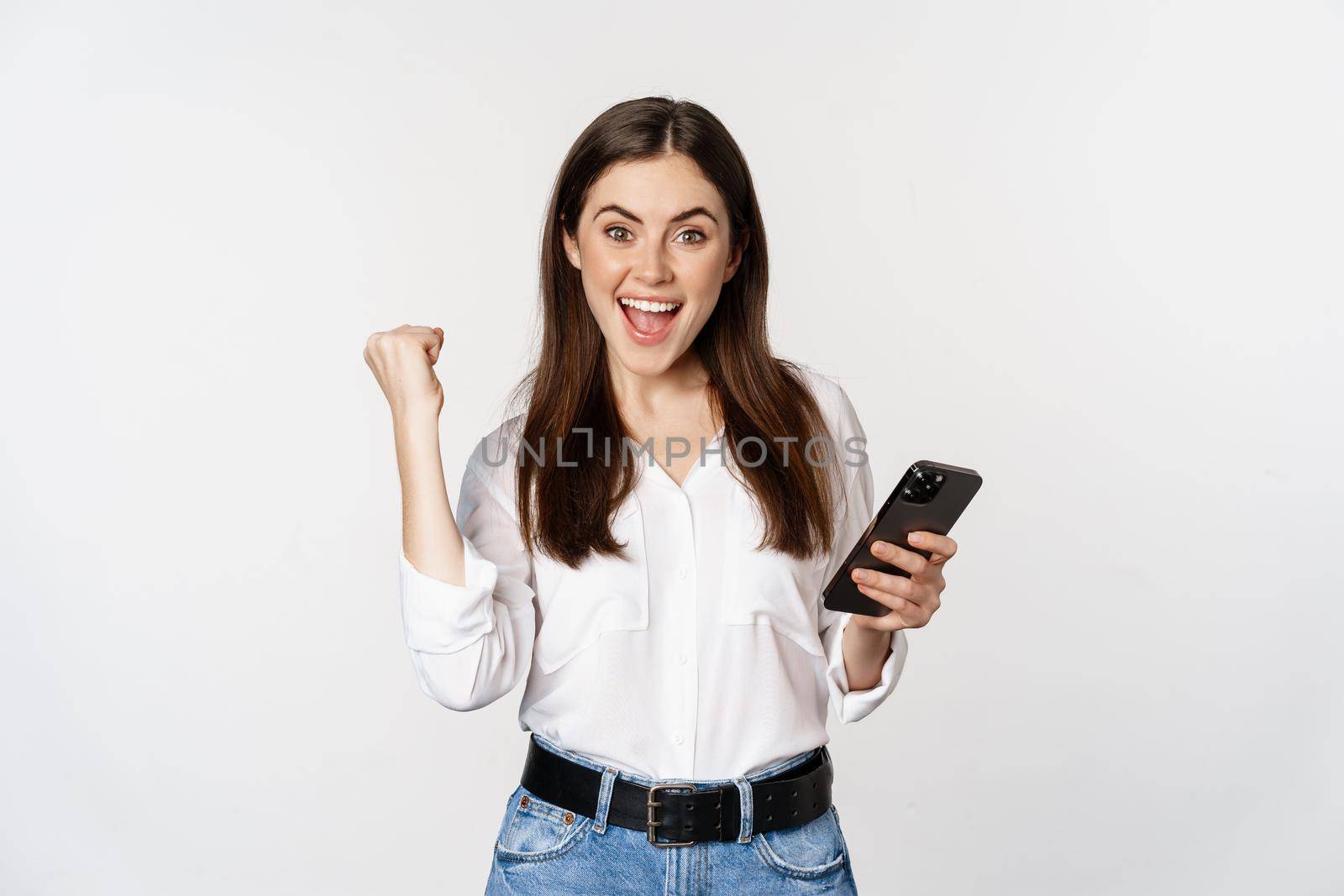 Enthusiastic woman winning on mobile phone, rejoicing and screaming of joy while using smartphone app, standing over white background.