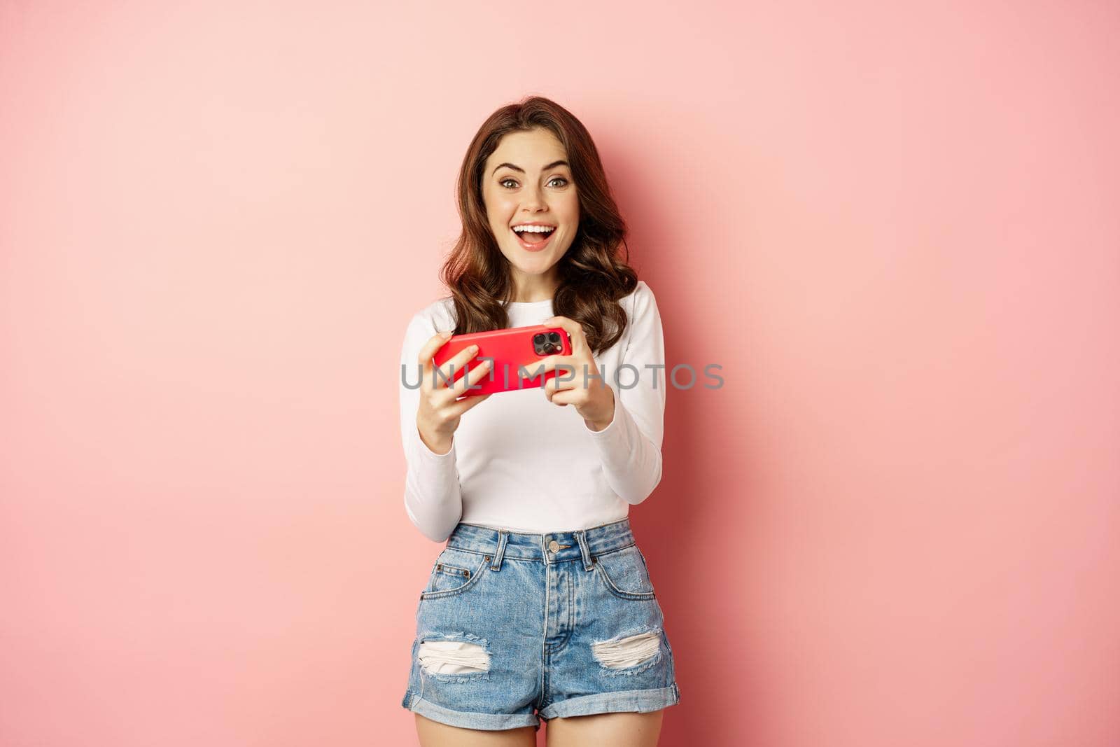 Happy beautiful girl laughing and smiling, watching video, playing games on mobile phone, horizontal smartphone, standing over pink background.