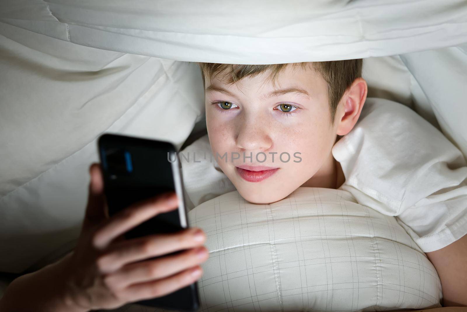 Boy under the blanket at night in his bed communicates on Internet. Child gadget addiction and insomnia. social media addiction by PhotoTime