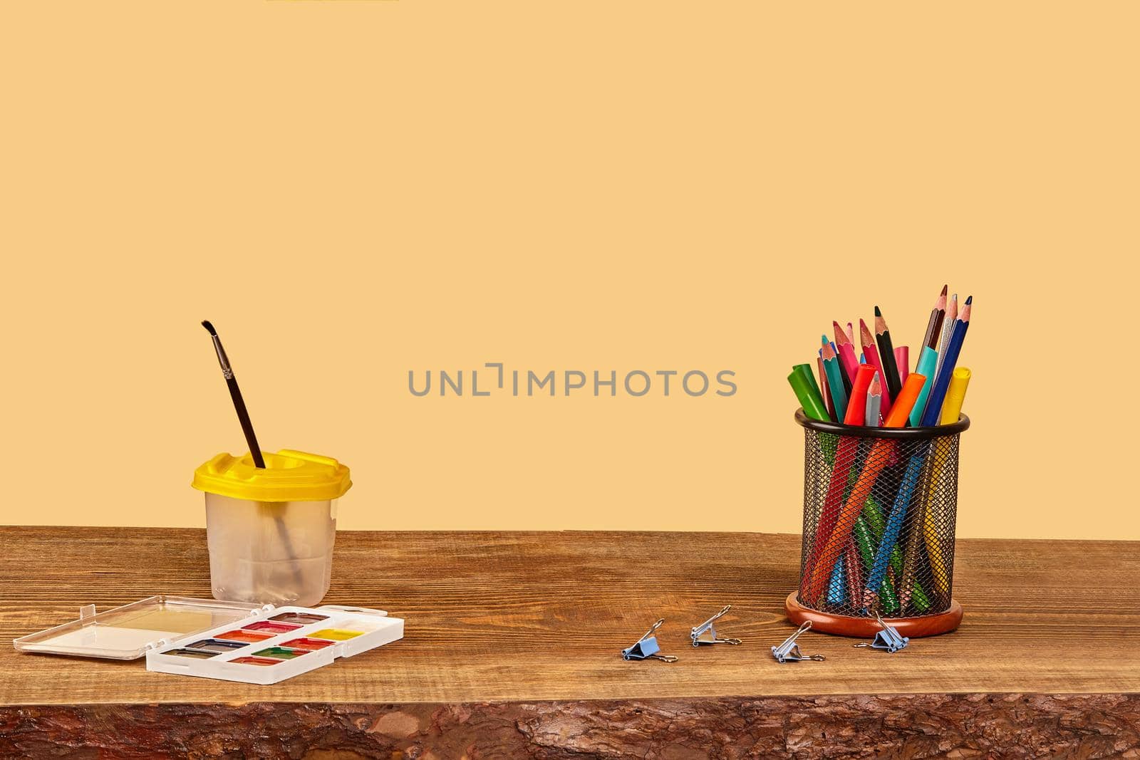 Watercolor set, paintbrush, binder clips, felt-tip pens and colorful pencils in black stand on a wooden desktop against beige background. Close up, copy space for your text or images. Mock up