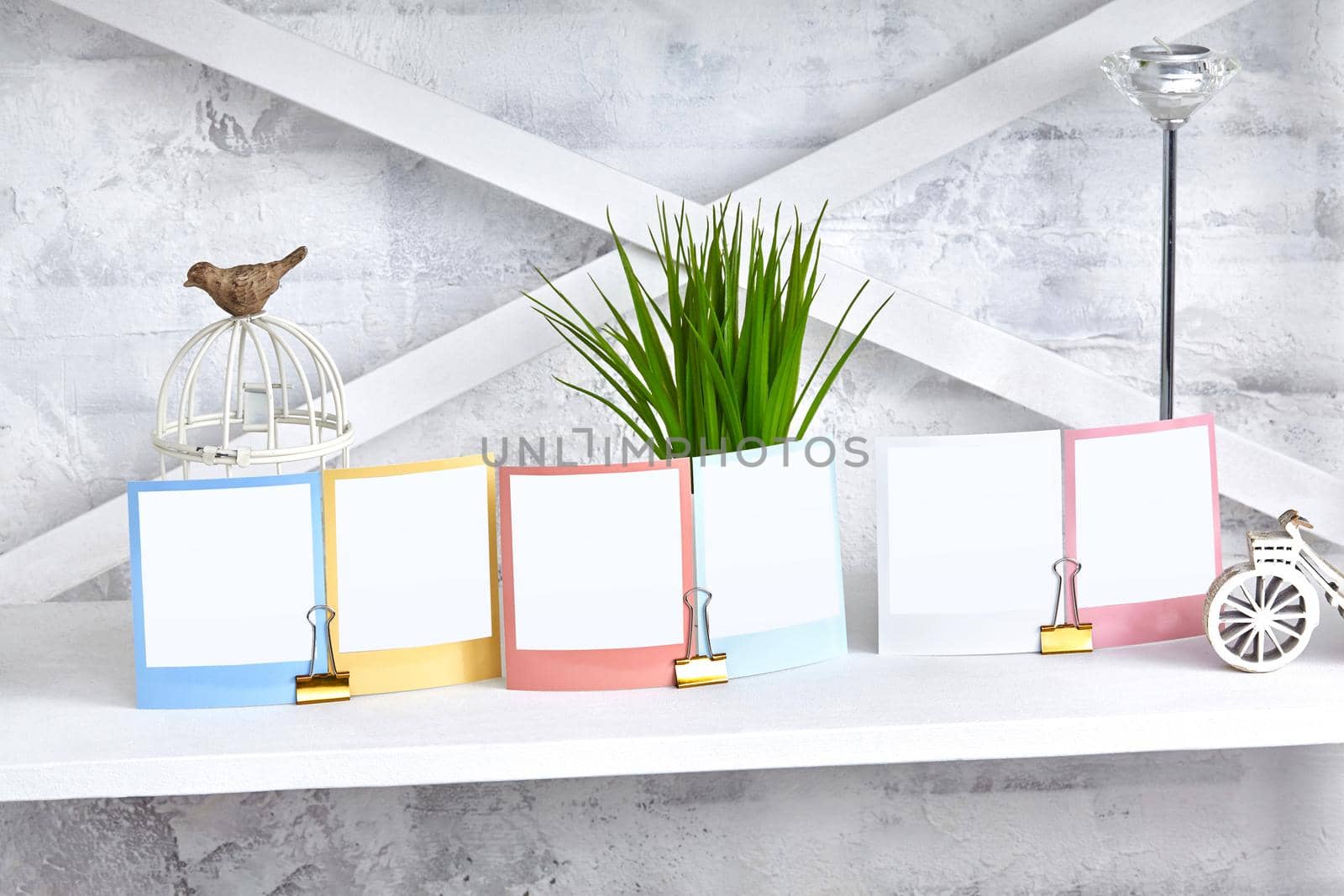 Colorful reminders, blank pieces of paper clamped by binder clips on white shelf, green plant and figurine of bird sitting on decorative cage, candle in silvery candlestick. Close up, copy space