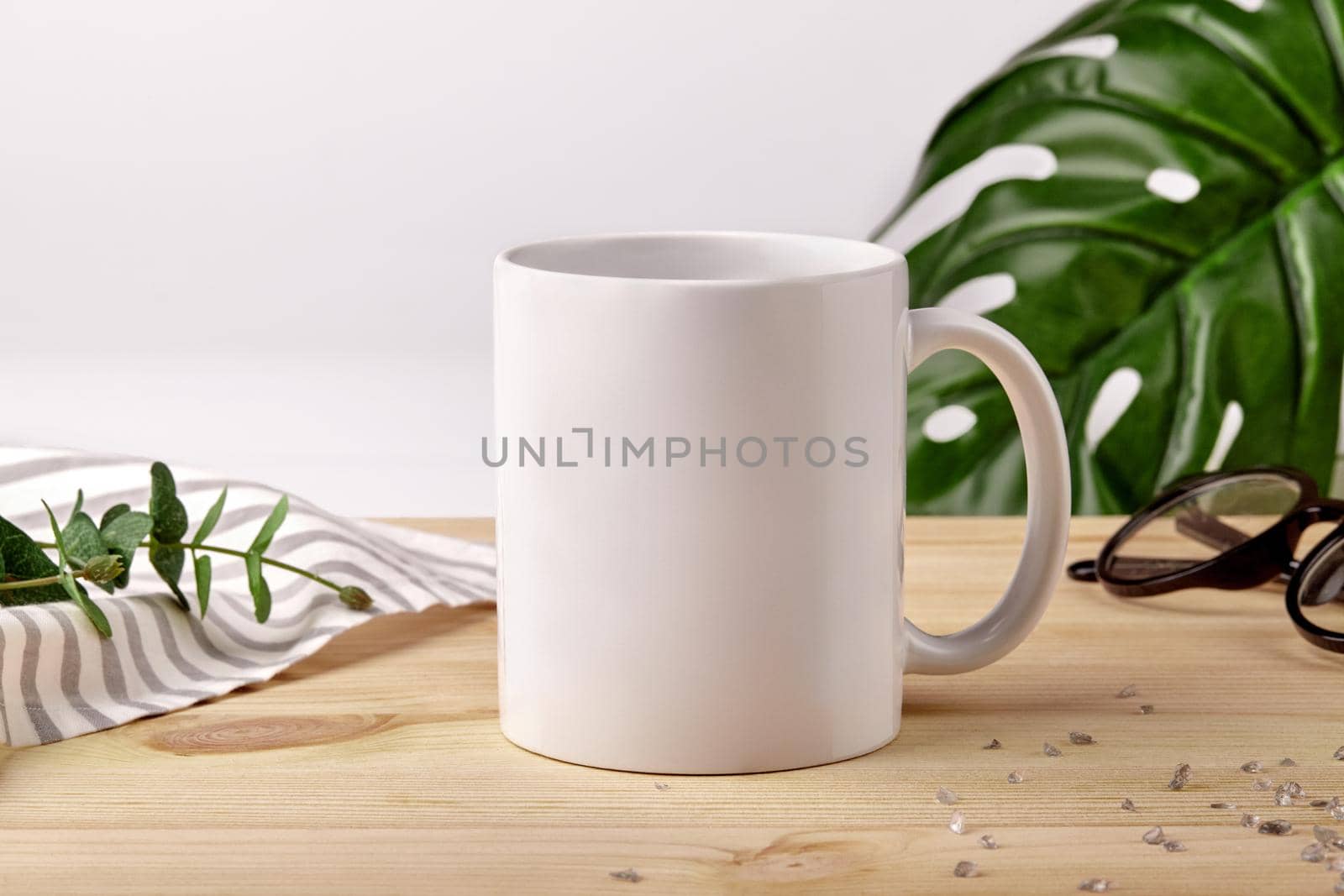 Ceramic mug for coffee or tea on wooden desktop next to striped tablecloth, scattered crystals and green plants against white studio background. Mock up, branding area. Close up, copy space