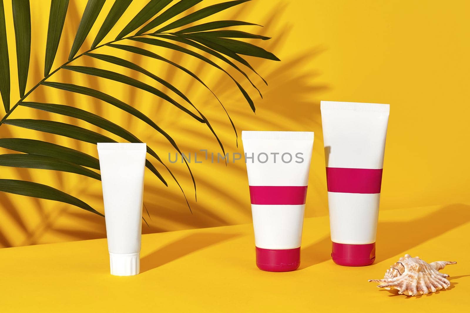 One white and two colorful cosmetic tubes with no logo next to a shell and green palm twig on yellow studio background. Close up, copy space by nazarovsergey