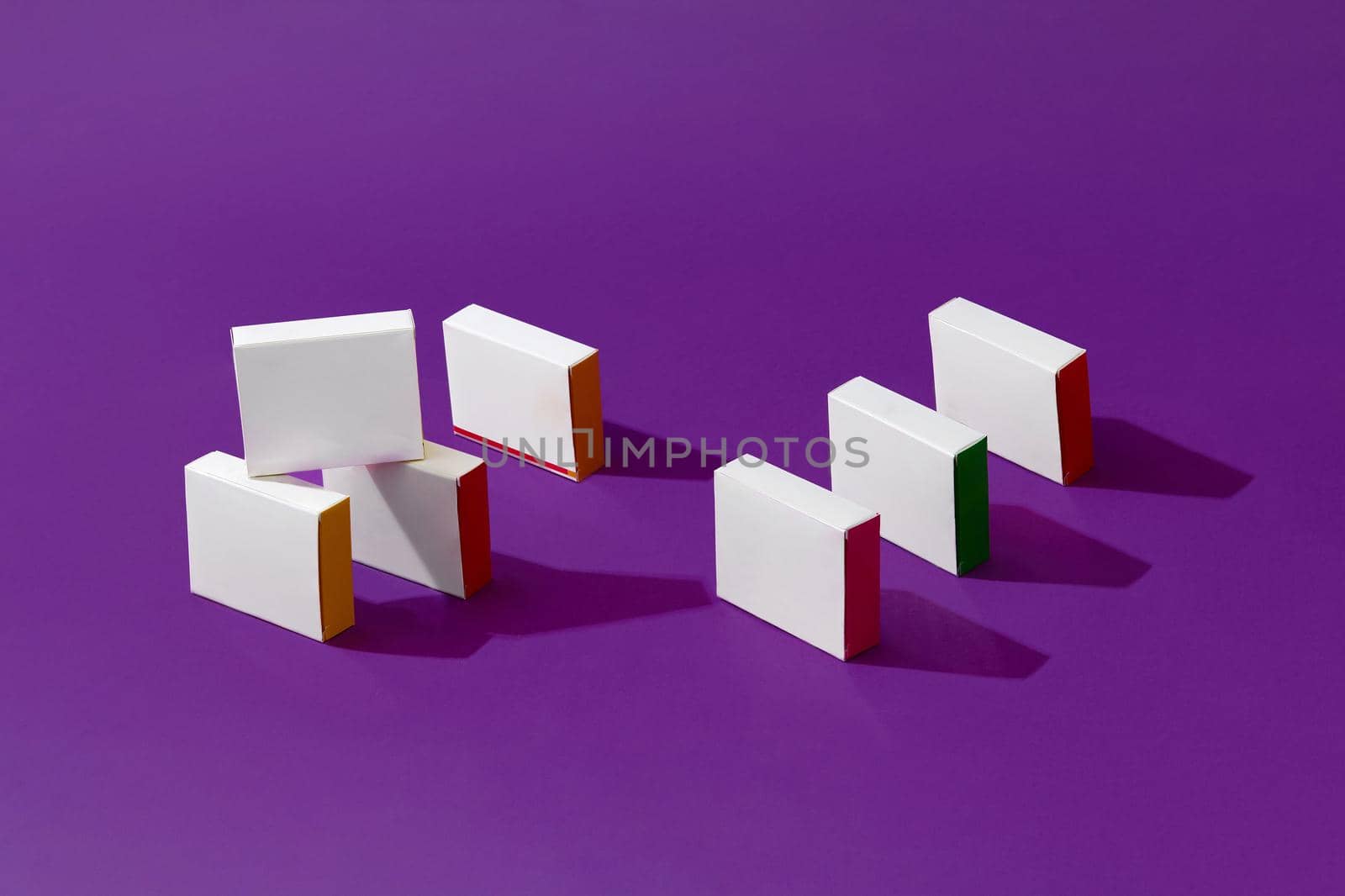 Seven small shadow-casting white cardboard boxes or packs with colorful sides and no logo on purple studio background. Branding area. Mock up, advertising. Close up, copy space