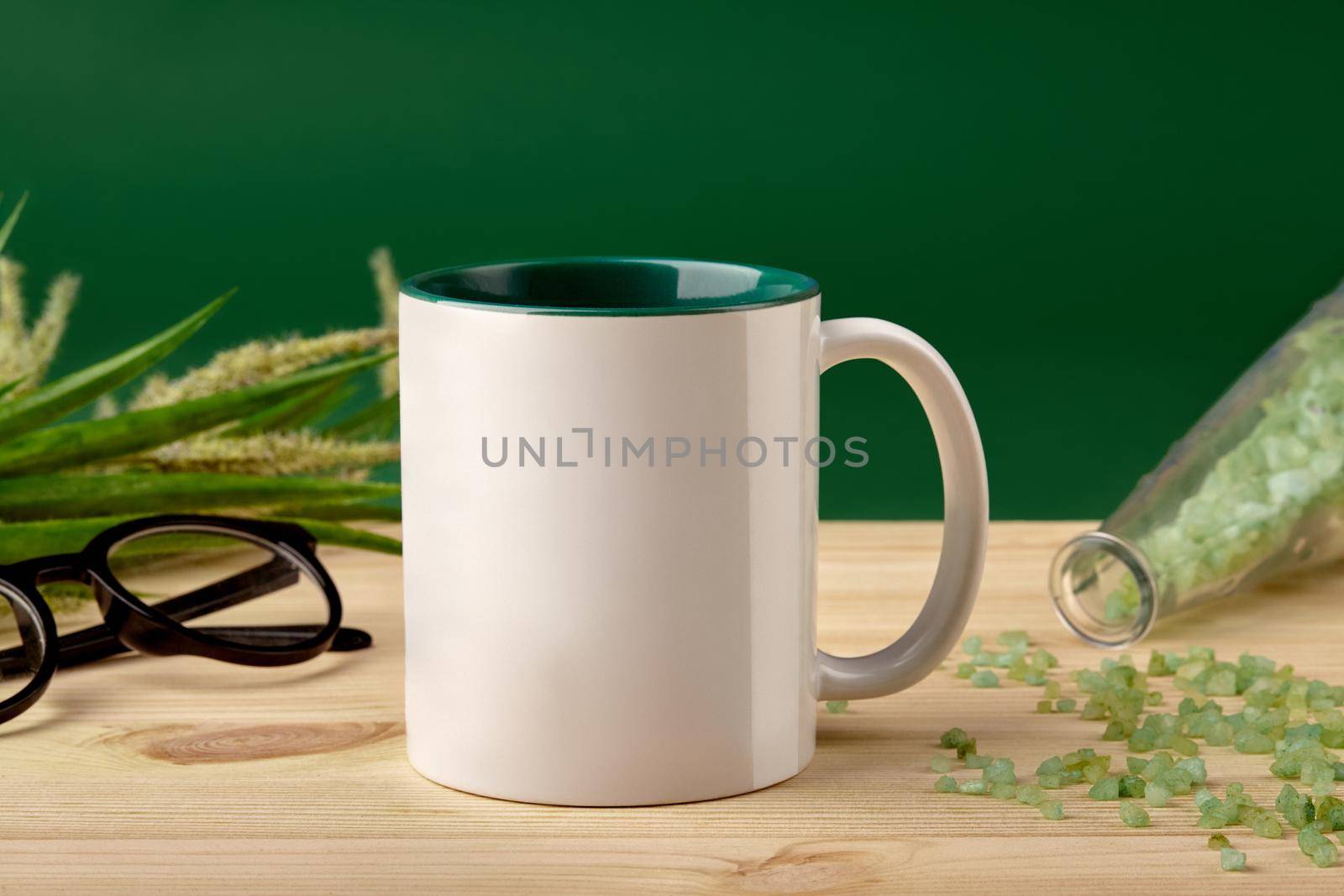 White ceramic mug, coarse sea salt scattered from the bottle on wooden desktop, glasses and blooming spikelets against green studio background. Mock up, branding area. Close up, copy space