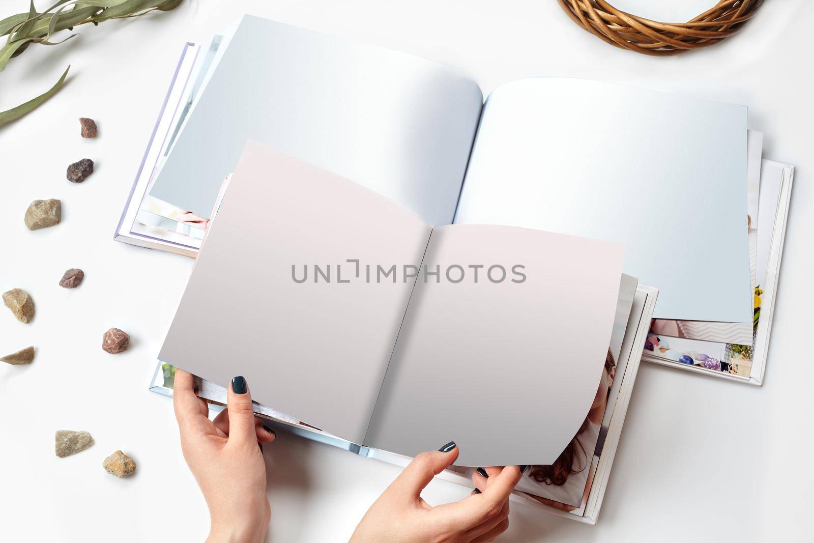 Hands of a female who holding two open photo books with hardback and blank pages, green twig and small stones isolated on white studio background. Close up, copy space. Mock up, top view, flat lay