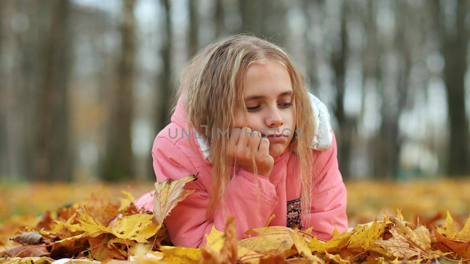 A little girl indifferently lies on the autumn foliage in a city park. by DovidPro