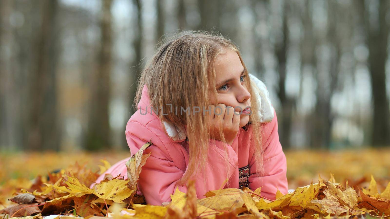 A little girl indifferently lies on the autumn foliage in a city park. She is very sad