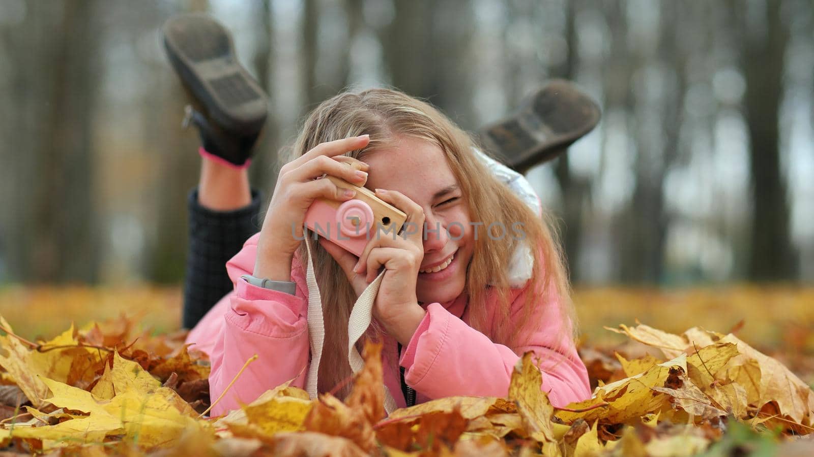 A teenage girl with a toy camera takes pictures of lying in the autumn foliage in the park