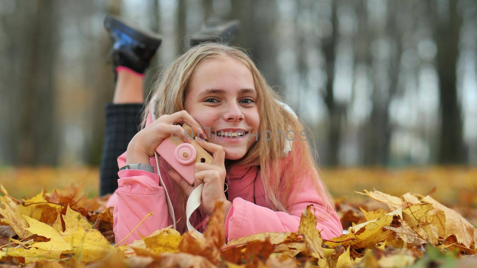 A teenage girl with a toy camera takes pictures of lying in the autumn foliage in the park. by DovidPro