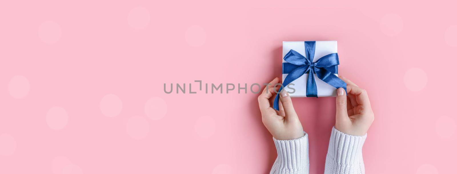 Banner of Female Hands in sweater holding gift in white wrapping paper on pink background. St. Valentines Day, love, tenderness, friendship, Birthday, Christmas concept. Festive wallpaper. Color 2022