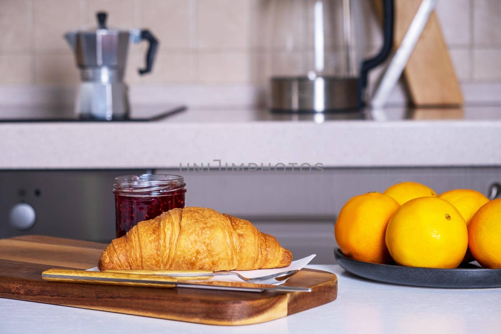 Wooden serving board with fresh croissant and jam and dish of oranges lies on kitchen table in a cozy kitchen. Selective focus.