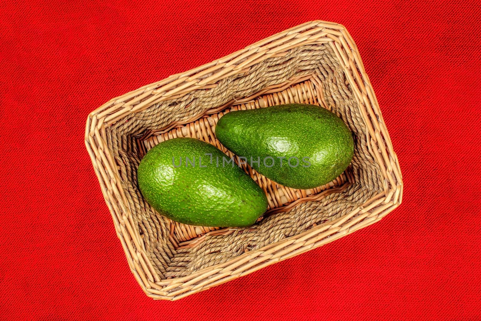 Table top view on two whole avocado fruits in basket, with red tablecloth under it.