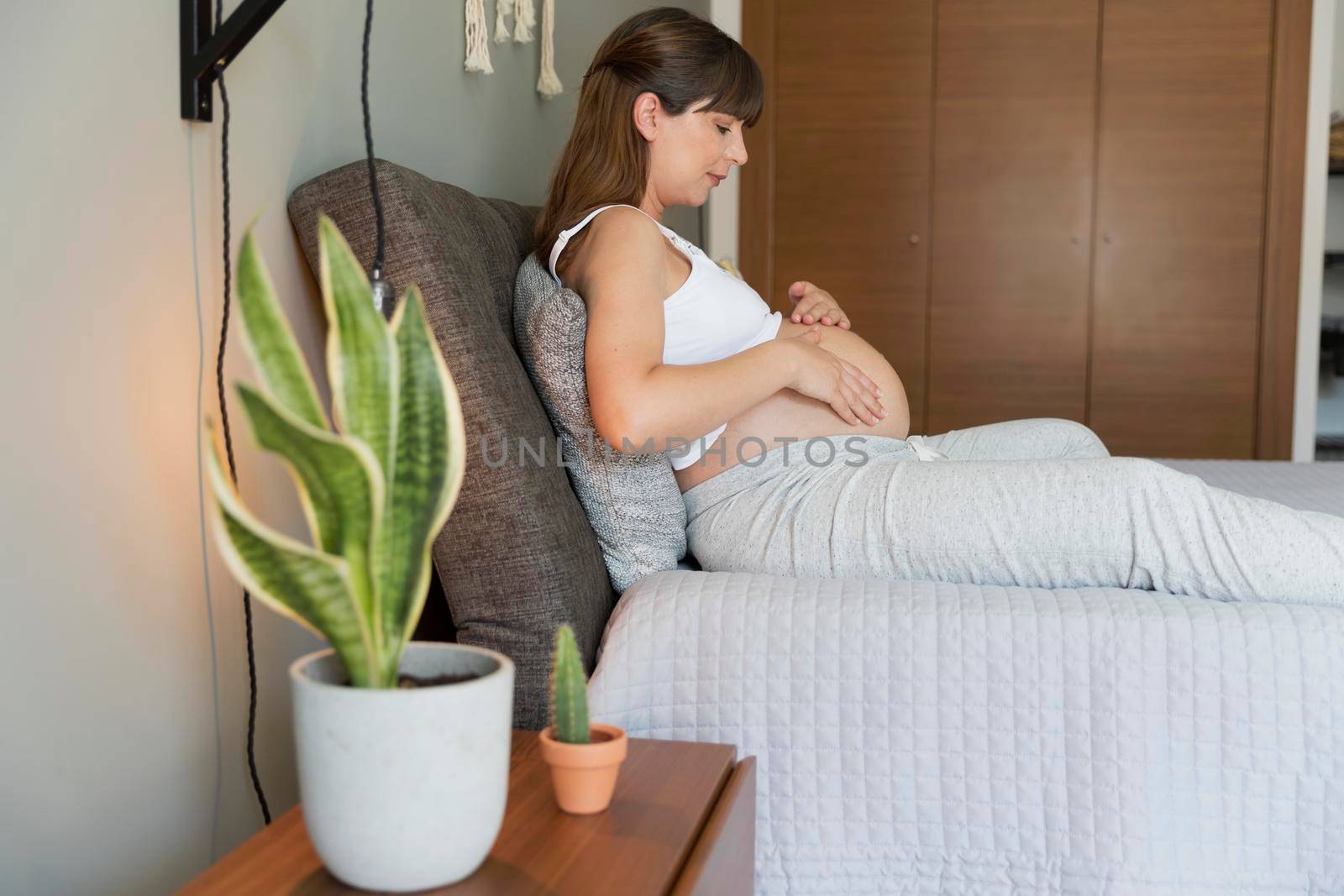 Happy pregnant woman sitting on bed and touching her belly at home