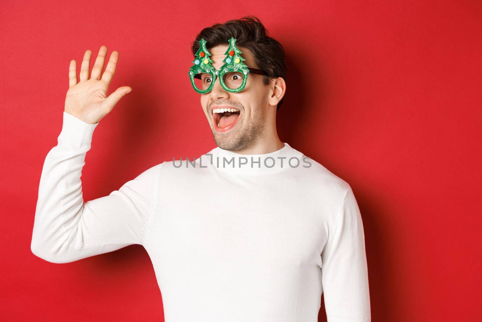 Portrait of friendly, cheerful man in party glasses and white sweater, saying hello and looking left, greeting a friend, standing over red background.