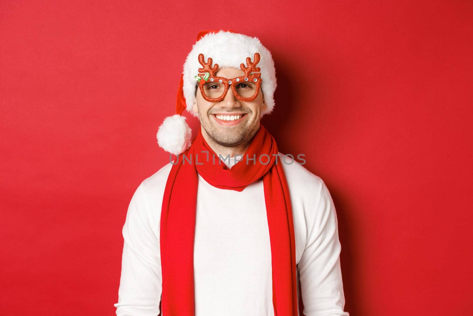 Concept of christmas, winter holidays and celebration. Cheerful handsome guy in new year party glasses and smiling, standing over red background.