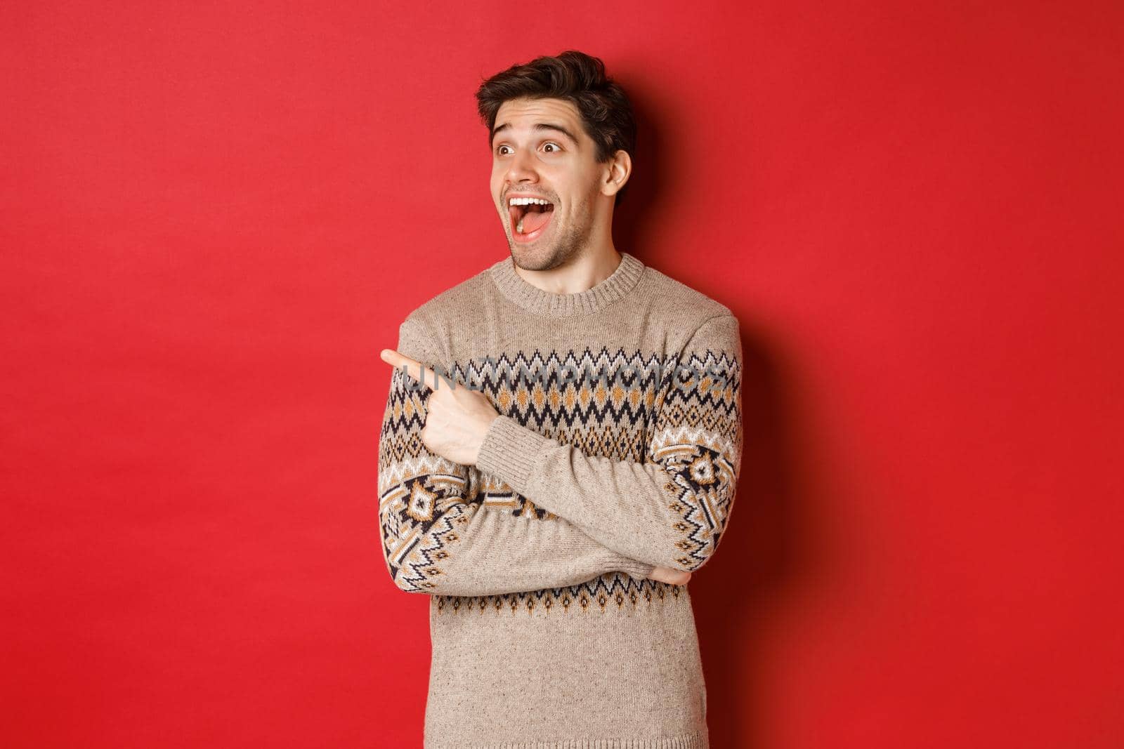 Portrait of attractive caucasian man celebrating new year, winter holidays, pointing finger and looking left with amazed expression, showing christmas advertisement, standing over red background.