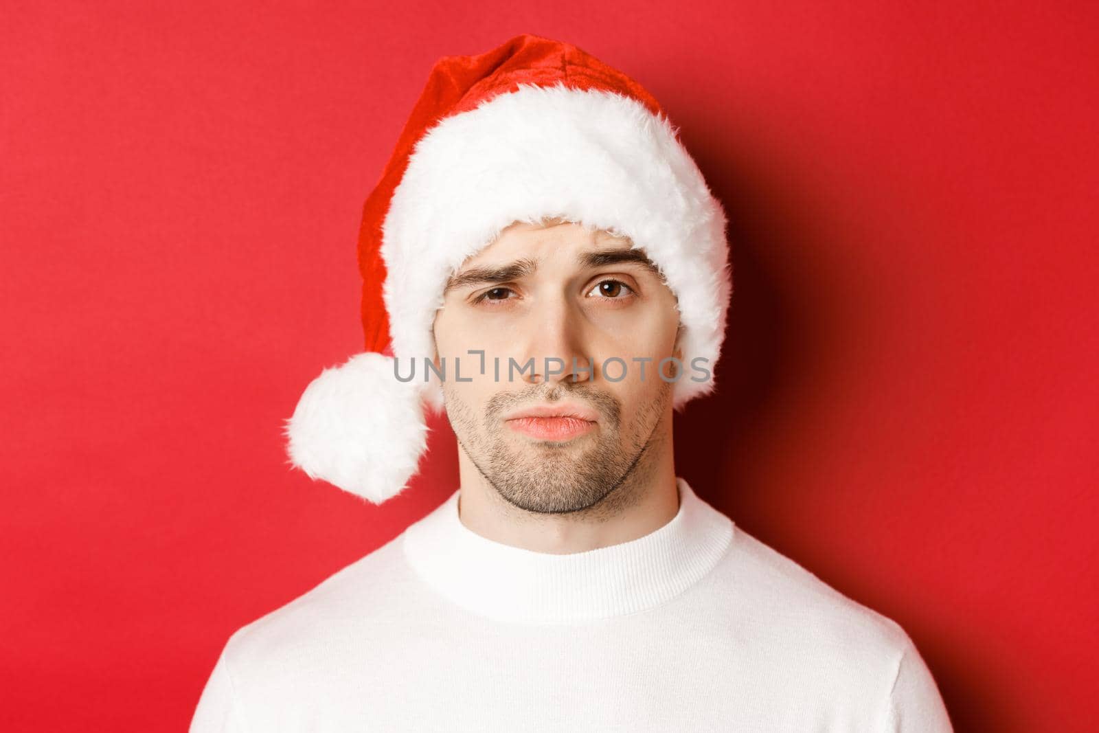 Close-up of doubtful frowning man, wearing santa hat and looking at camera uncertain, standing against red background.