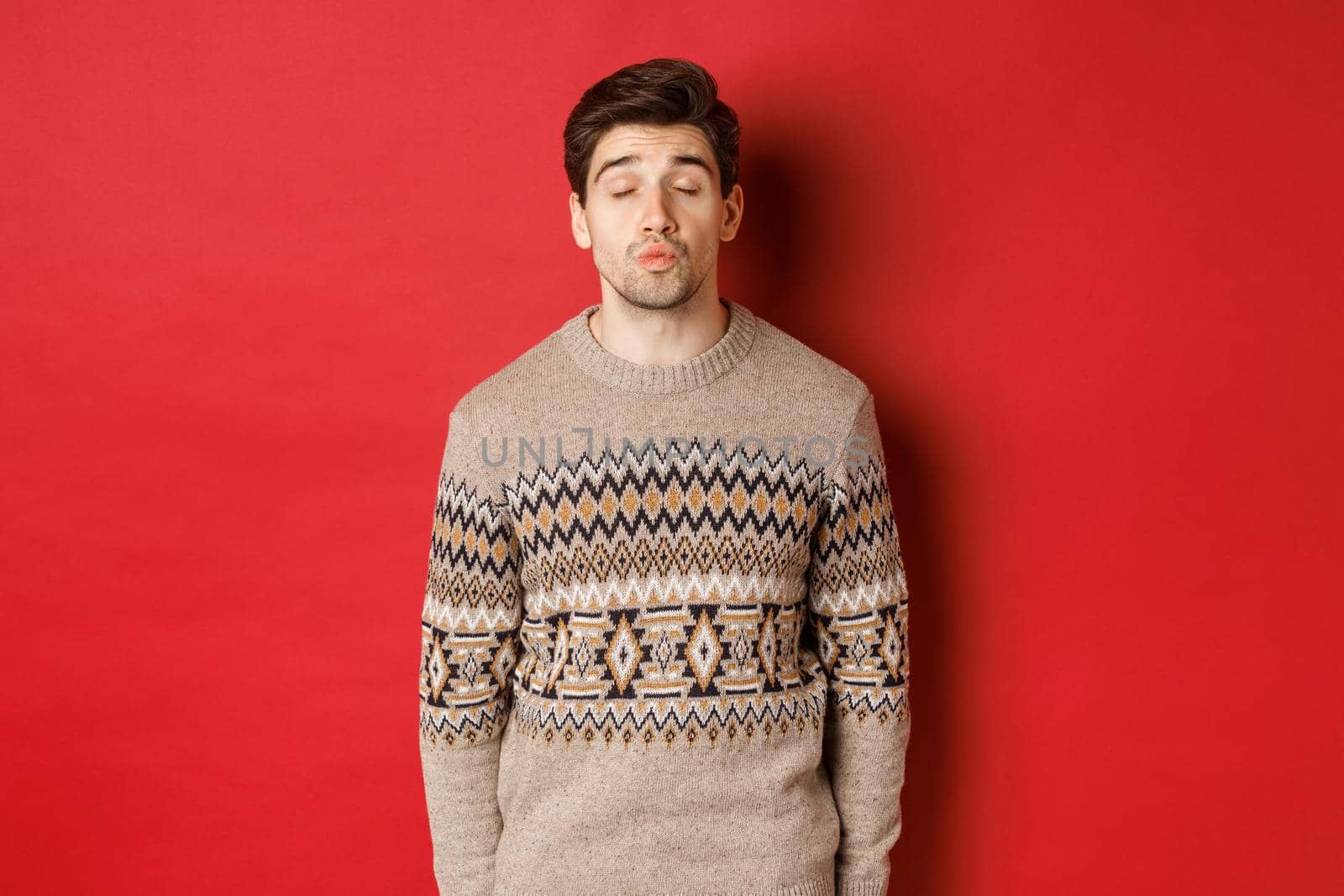 Image of handsome man in christmas sweater pucker lips and close eyes, waiting for kiss under mistletoe, standing over red background.