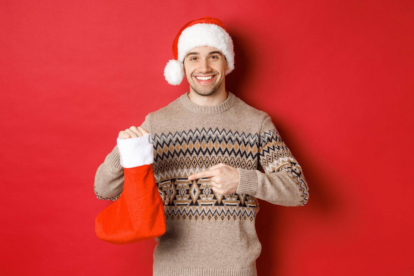 Concept of winter holidays, new year and celebration. Handsome smiling man prepared gifts for kids, pointing at christmas stocking bag, standing over red background.