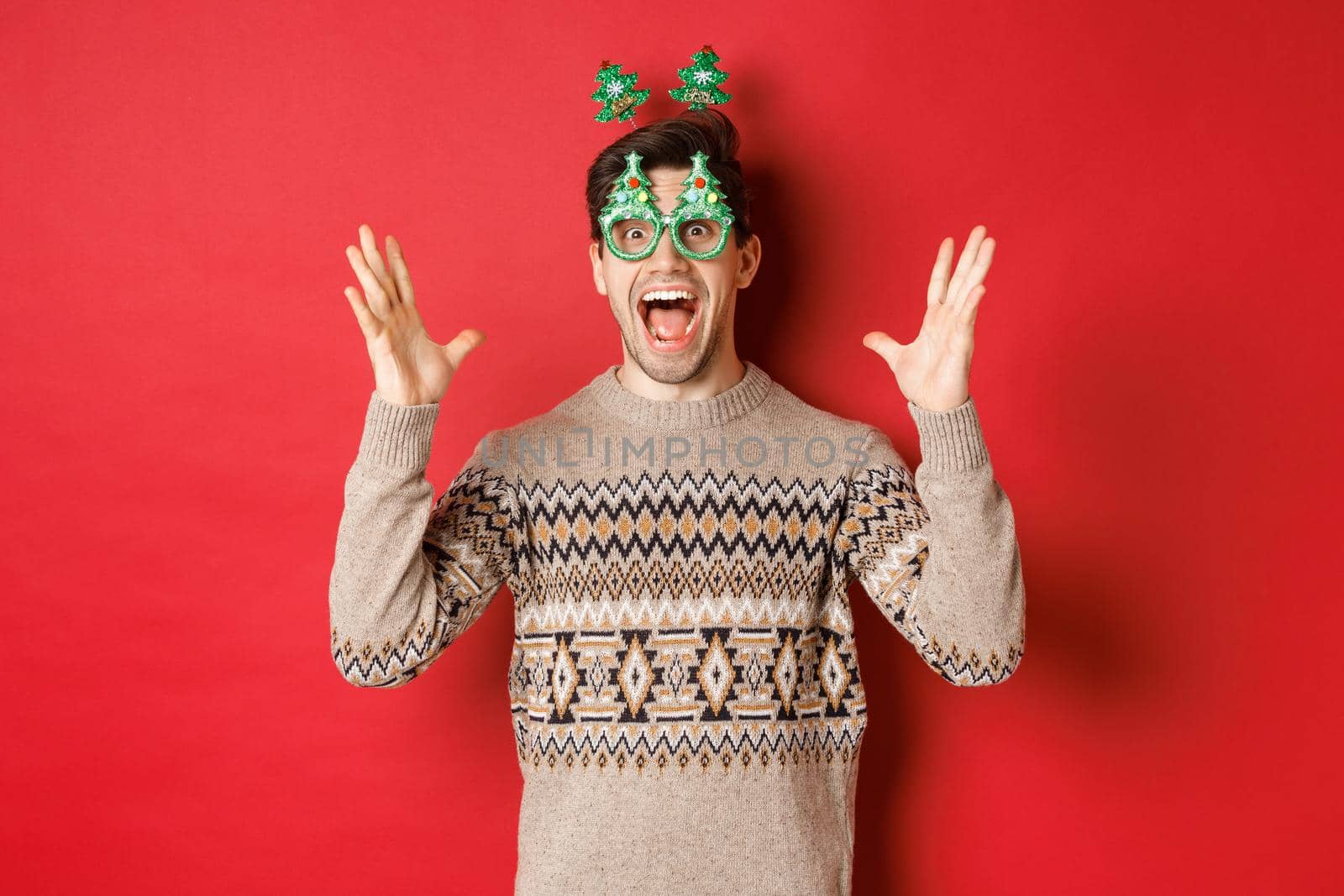 Image of excited and amazed caucasian guy in party glasses, christmas sweater, raising hands up and making big announcement, enjoying new year celebration, red background.