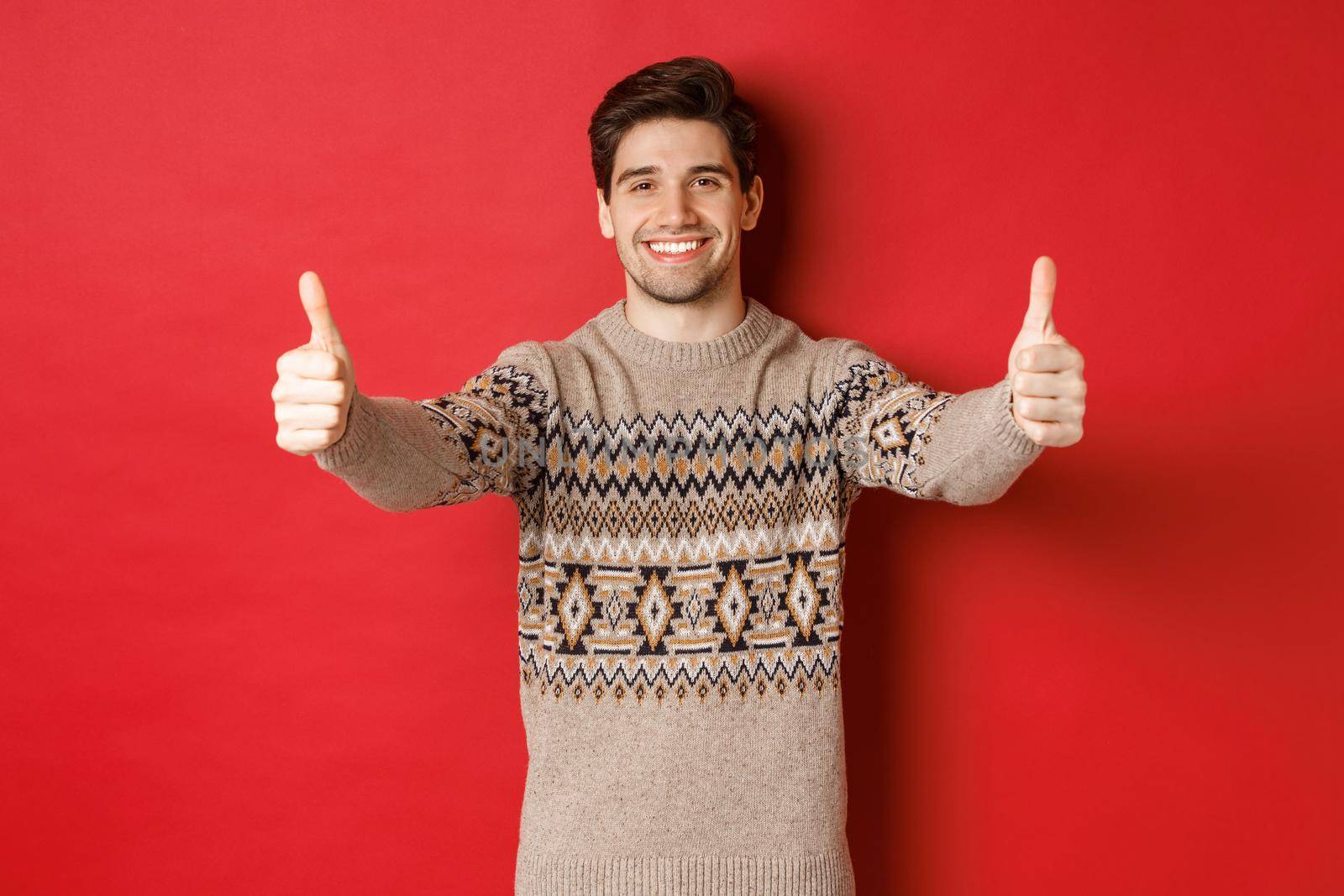 Image of handsome caucasian man in christmas sweater, showing thumbs-up in approval and smiling, wishing happy holidays, standing over red background.