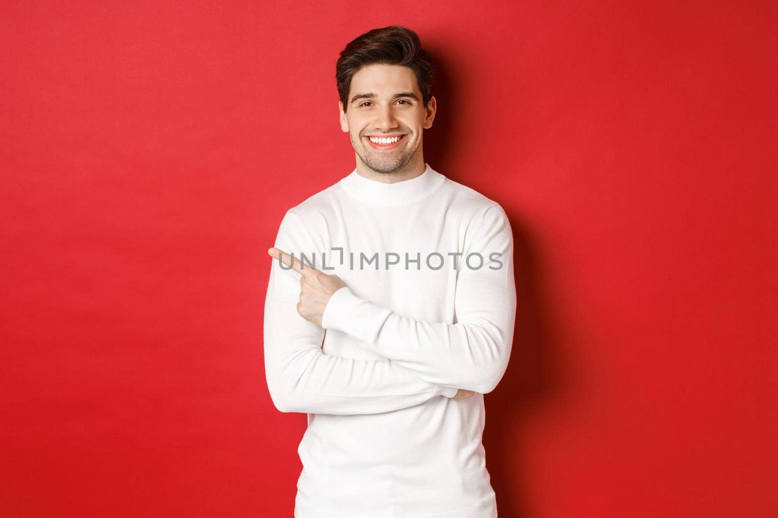 Concept of winter holidays, christmas and lifestyle. Portrait of attractive man in white sweater, pointing finger at upper left corner and smiling, showing logo on red background.