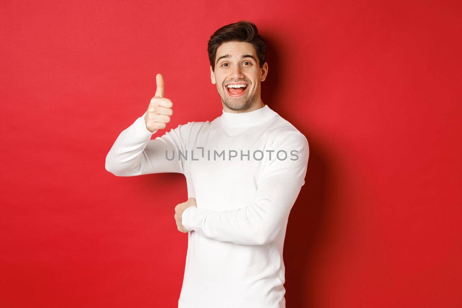 Concept of winter holidays, christmas and lifestyle. Portrait of cheerful, attractive man in white sweater, showing thumbs-up in approval, like good idea, standing over red background.