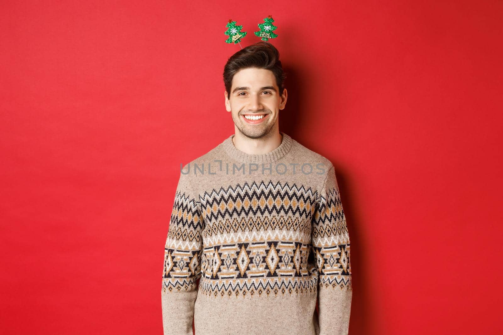 Concept of winter holidays, christmas and celebration. Handsome bearded guy in sweater wishing happy new year, smiling at camera, standing over red background.