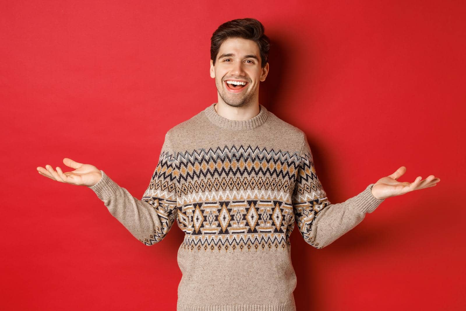 Portrait of happy good-looking man celebrating new year holidays, wearing christmas sweater, spread hands sideways and smiling, holding something on copy space, standing over red background.